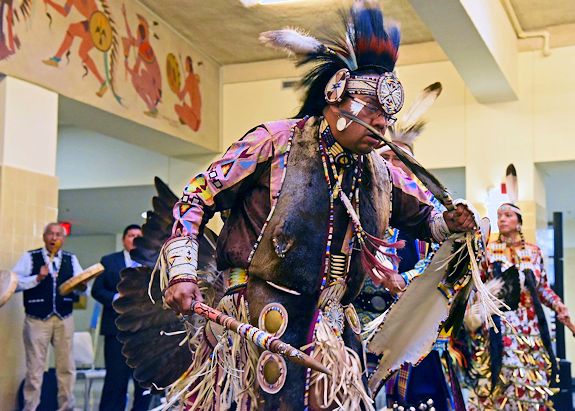 A Native American man wearing traditional clothes and feathers dances inside a large room.